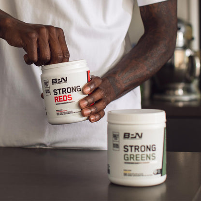Person holding a container of BSN Strong Reds supplement with another container of BSN Strong Greens on a surface.